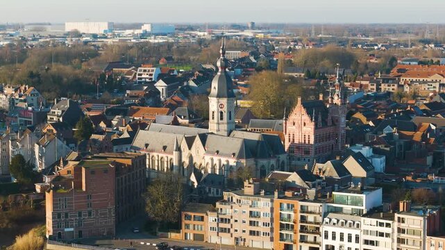 sharp grand stone church domed clock tower above dense belgian rooftops alongside ornate red brick historic building among modern apartments leafy streets up close aerial temse belgium 