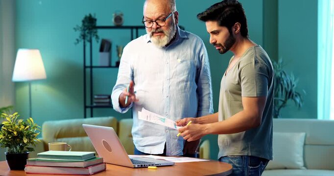 Indian young son and senior father filing online tax return together using laptop while standing at table in modern home, discussing financial reports, income statements yearly accounting documents