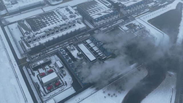 Aerial view of a large industrial data center complex covered in snow. Steam rises from the cooling towers on a cold, overcast winter day