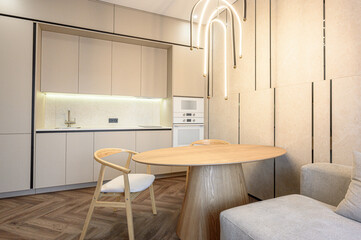 Fototapeta premium A modern kitchen with a round wooden table, light wooden chairs, built-in appliances, neutral cabinetry, and a stylish suspended LED light fixture — minimalist and cozy