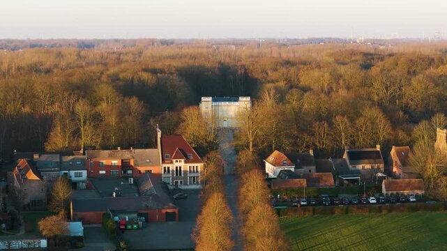 symmetrical golden hour bornem tree lined avenue leading pale yellow fa&ccedil;ade d&rsquo;ursel castle framed by warm lit village houses autumn toned woodland to drone shot belgium 