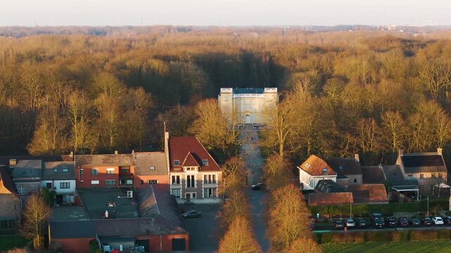 closer golden hour perspective bornem warm lit village rooftops townhouse sun bathed yellow d&rsquo;ursel castle emerging through leafless trees forest's edge close drone belgium stately 