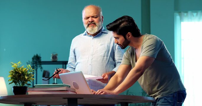 Indian young son and senior father filing online tax return together using laptop while standing at table in modern home, discussing financial reports, income statements yearly accounting documents