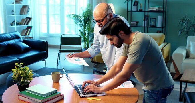 Indian young son and senior father filing online tax return together using laptop while standing at table in modern home, discussing financial reports, income statements yearly accounting documents