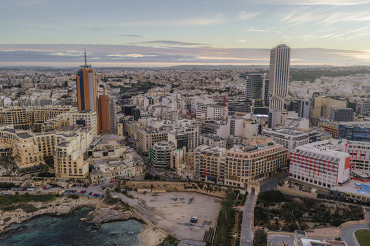 Aerial view of buildings casting long shadows under a soft, diffused light, with the sea's turquoise waters contrasting against the city's pale stone, St. Julian's, Malta.