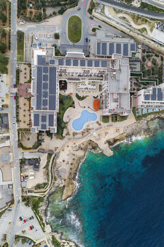 Aerial view of the azure waters crashing against the rugged coastline, juxtaposed with the geometric architecture and pool of the Radisson Blu hotel, St. Julian's, Malta.