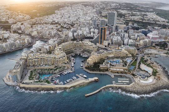 Aerial view of Portomaso Marina's yachts nestled amidst the upscale hotels and gleaming towers under the soft, golden light, St. Julian's, Malta.
