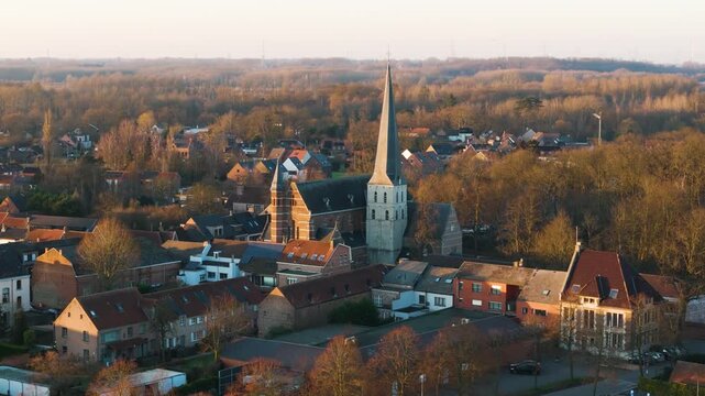 warm golden hour bornem focuses tall stone church spire above tightly packed red roofed houses soft evening light village forested hills up close aerial belgium red-roofed 