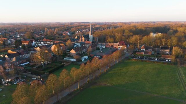 golden hour bornem depicts rows warm lit village houses clustered tall church spire bordered by tree lined road green fields fading into glowing forest around drone view belgium 