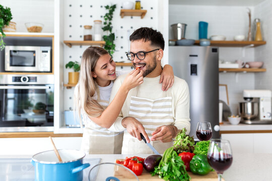 Happy Young Couple Tasting Food while Cooking. Fun in the Kitchen, Food and Drink concept. Attractive woman feeding man a piece of vegetable. People preparing healthy meal together at home.