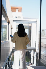 Fototapeta premium Woman standing by window holding phone working remotely