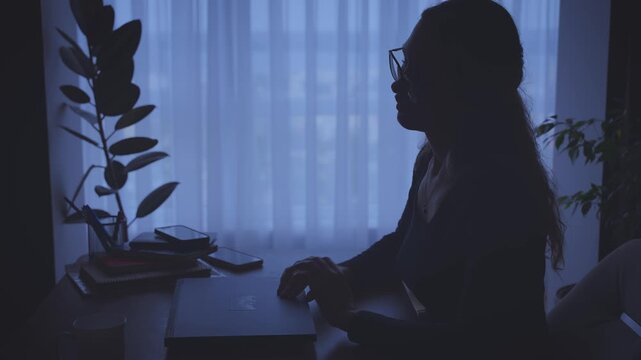 dark silhouette unhappy tired woman working on laptop overloaded with work, sitting at desk in office. Sad face girl suffers from headache. depression fatigue lack of money. Person looking for job.