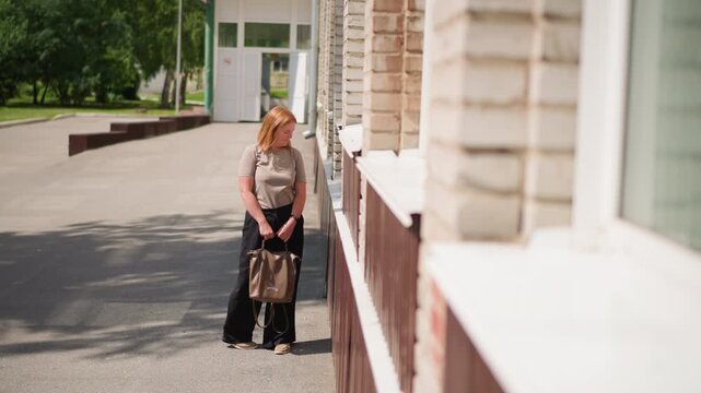 Young woman holding bag walking toward brick building wall under bright sunlight, preparing to lean and rest, thoughtful expression in quiet outdoor scene, symbolizing contemplation
