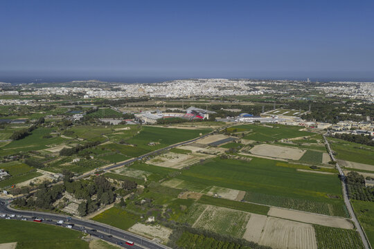 Aerial view of lush green fields stretching towards the distant city under a clear sky, a vibrant tapestry of earth and civilization, Rabat, Mdina, Malta.
