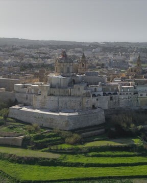 Aerial view of the ancient walled city gleams under the sunlight, with its honey-colored stone buildings and verdant terraced fields creating a striking contrast, Rabat, Mdina, Malta.