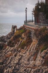 A winding pathway hugs the rocky shoreline as waves crash below. Street lamps stand tall, illuminating the natural beauty. The sky hints at rain, creating a moody yet tranquil atmosphere