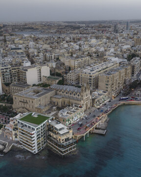 Aerial view of buildings lining the coast with the deep blue sea gently lapping at the shore, contrasting with the urban landscape, Sliema, Sliema, Malta.