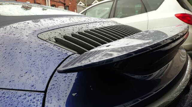 tight rear quarter blue porsche sports car rain dotted engine cover horizontal cooling slats raised spoiler against muted urban parking backdrop view paint decklid 
