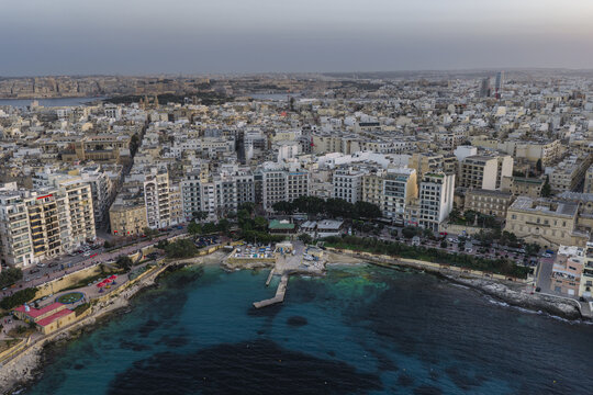 Aerial view of turquoise waters meeting the stark, blocky architecture of the city in a stunning contrast of nature and urban sprawl, Sliema, Sliema, Malta.