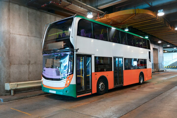 A contemporary double-decker bus in white, orange, and green sits stationary within a bright indoor public transport facility. Concrete walls and ceiling lights illuminate the spacious depot