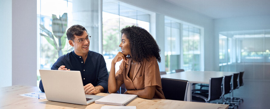 Happy busy African business woman and Hispanic business man talking in office working together on laptop computer at meeting. Two coworkers discussing corporate marketing project sharing ideas at work