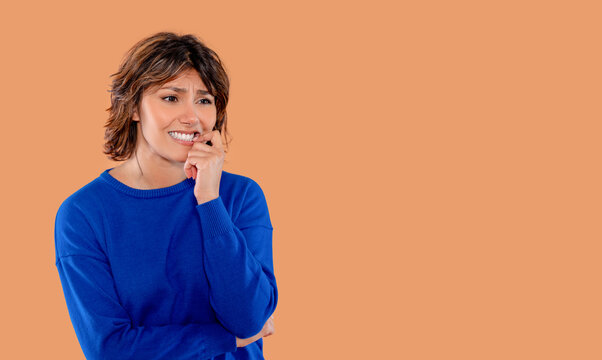 Woman looks worried while biting her nails in front of an orange background in a bright room