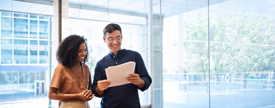 Business team of two happy young busy diverse colleagues, professional business man and business woman executives working together standing in office looking at project documents and talking.