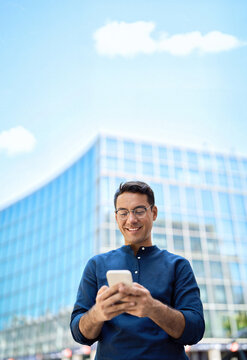 Happy professional businessman holding smartphone in hands looking at cellphone outdoor, young Hispanic business man customer using finance banking mobile app on cell phone standing in city. Vertical.