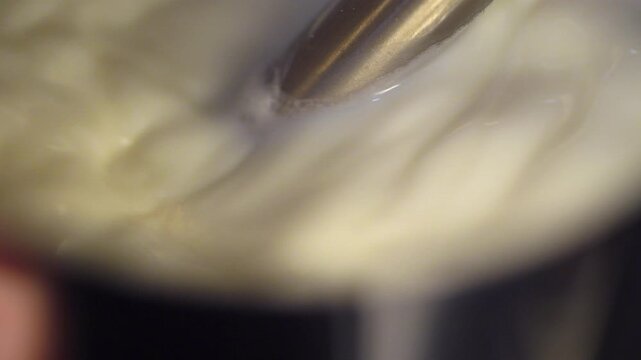 Extreme slow motion macro shot of milk being textured with a steam wand, creating glossy microfoam inside a stainless steel pitcher. Focus on fluid motion, heat, and creamy structure during espresso p