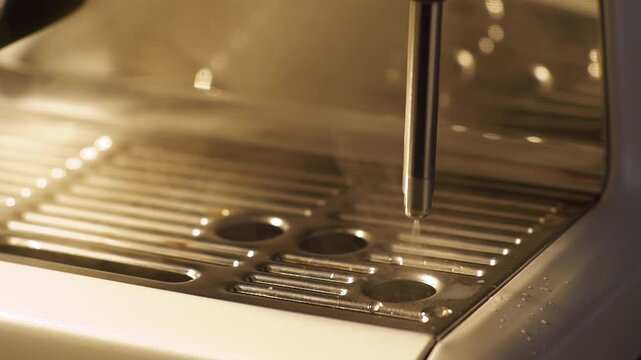 Close up slow motion shot of an espresso machine steam wand releasing hot vapor above the metal drip tray. Warm light reflections and rising steam highlight barista workflow and milk frothing preparat