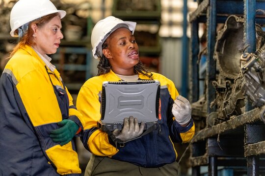 Two female engineers in safety gear inspecting car parts inventory. workers technical walking old dirty car industry checking used auto parts by tablet industrial automotive parts warehouse Recycling