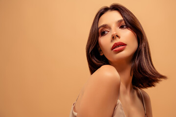 Pretty young woman with stylish brunette hair and flawless makeup, posing against a beige background
