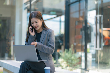 Fototapeta premium Businesswoman talks on phone, updates tasks, and plans milestones on a laptop. Online business, technology, in front of a modern office building.