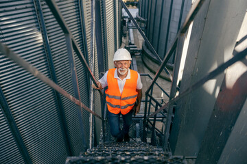 Senior white male worker wearing orange safety vest and hard hat standing outdoors at industrial facility