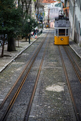 Naklejka premium LISBON, Portugal - MARCH 2025É Yellow tram on a street with colorful houses and flowers on the balconies - Bica Elevator going down the hill of Chiado
