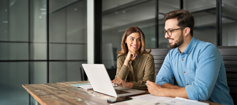 Two happy professional business people, smiling colleagues or company workers talking at meeting using laptop computer discussing online technology project together working in office at workplace.