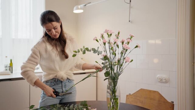 Caucasian daughter standing in kitchen. Young female placing flowers into glass vase. Smiling woman arranging bouquet with care. Enjoying family tradition at home. Daytime interior scene.