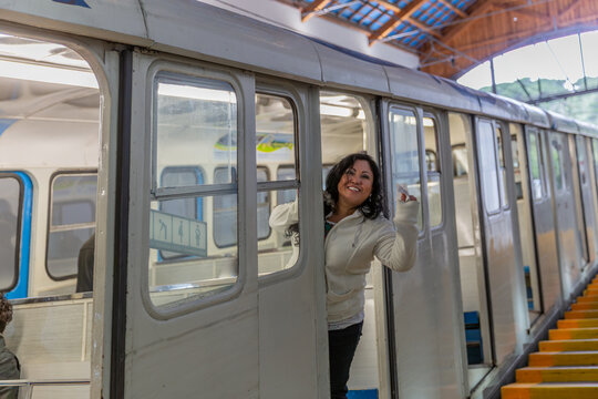 Smiling and happy middle-aged adult female tourist looking out of Tibidabo funicular cabin, white metal car with multiple windows, rails on slope of Cuca de Llum station in Barcelona, Spain