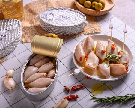 Gourmet canned small squids in olive oil with rosemary and chili peppers on a white plate, tapas