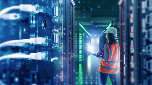 Data center operations, IT maintenance. Engineer in safety vest monitors server racks with laptop, checking network equipment, data storage systems and cybersecurity infrastructure.
