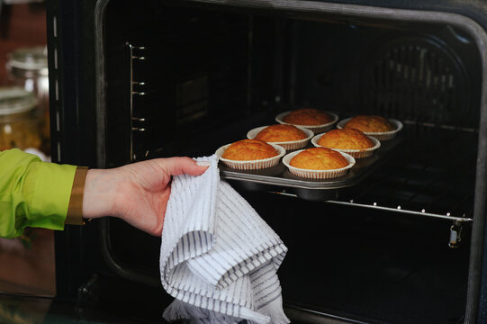 A close-up shot of a person's hand using a white striped kitchen towel to pull a metal baking pan with six golden-brown muffins out of a dark oven. The cupcakes are perfectly baked in white paper line