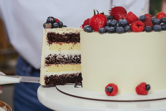 A professional close-up shot showing a perfect slice being taken from a tall white frosted cake. The cut reveals distinct, moist layers of dark chocolate sponge and light vanilla cream filling. The to