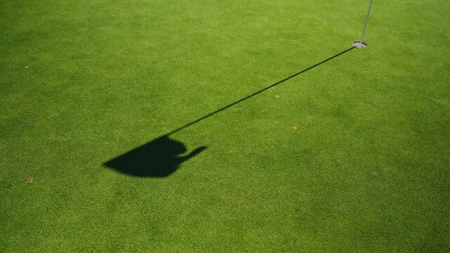Long shadow of a flagstick stretches across a manicured green as it waves in the wind