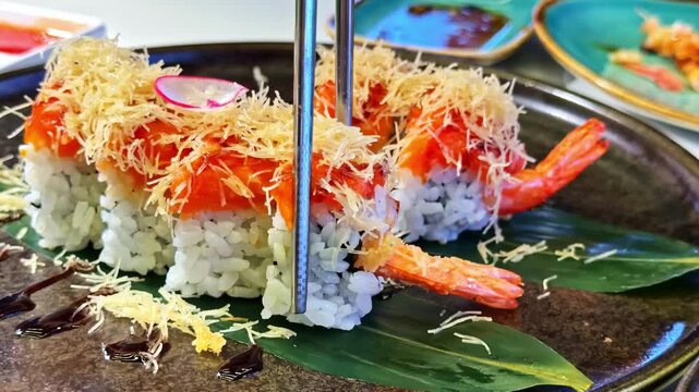 Close up footage of an ebi tempura sushi roll lifted with chopsticks at a restaurant table. Crispy shrimp, sushi rice, and sauce topping highlight fresh Japanese dining.