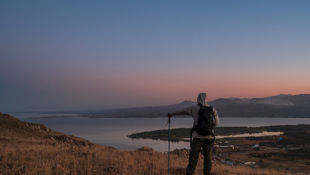 A hiker wearing a hooded jacket and backpack stands on a hill overlooking Lake Sevan at dusk.
