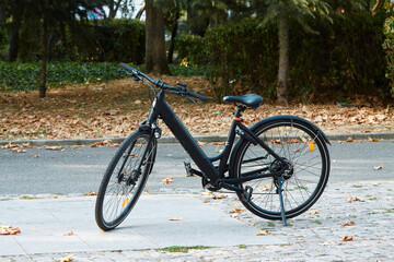 Fototapeta premium Black electric bicycle parked on a paved surface with fallen leaves and trees in the background during autumn season