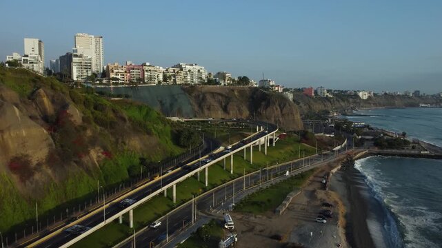 Aerial footage of a coastal highway called "Costa Verde". Drone then pans left showing a road called "Subida Armendariz" which has a blue bridge called "Puente de la paz". Located in Lima, Peru.