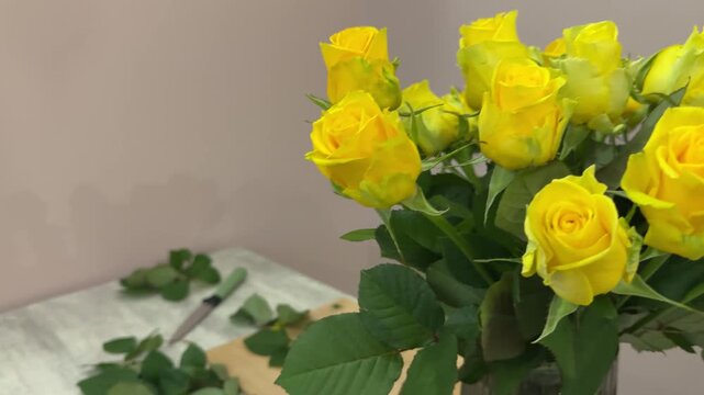 Luxurious bouquet of yellow roses with messy florist workspace, leaves, thorns and cut stems on table, authentic floral design process on neutral background, behind the scenes concept.