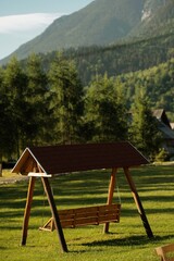 A rustic wooden swing rests beneath a charming shelter in a peaceful meadow. The backdrop features majestic mountains and vibrant green trees, creating a tranquil escape
