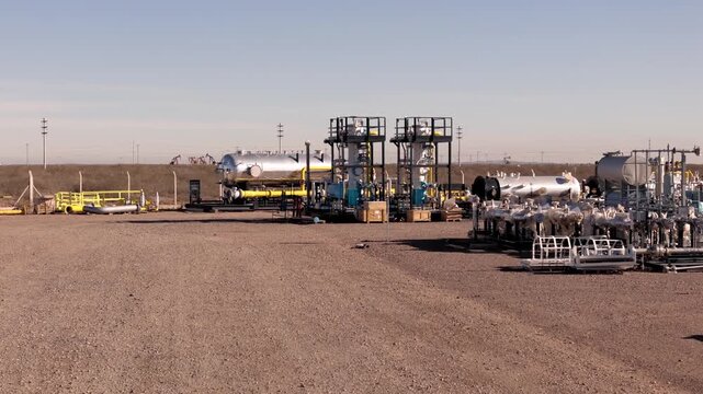 Drone footage moving closer to surface processing modules and storage tanks at an onshore oil and gas production facility in an arid industrial landscape.